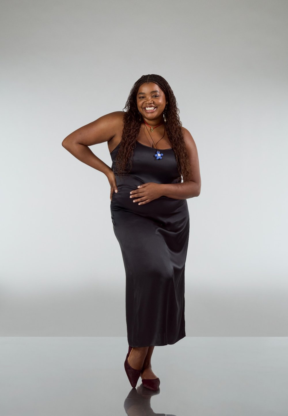 Černé saténové šaty na ramínka s přiléhavým střihem. Modelka má na sobě modrý přívěsek ve tvaru květu a bordó podpatky, stojí na neutrálním pozadí.