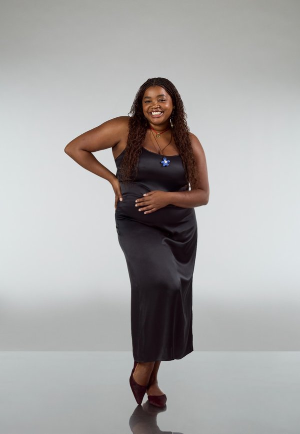 Zwarte satijnen slipjurk met een aansluitende silhouet. Het model draagt een blauw bloemensieraad en bordeauxrode hakken en staat tegen een neutrale achtergrond.