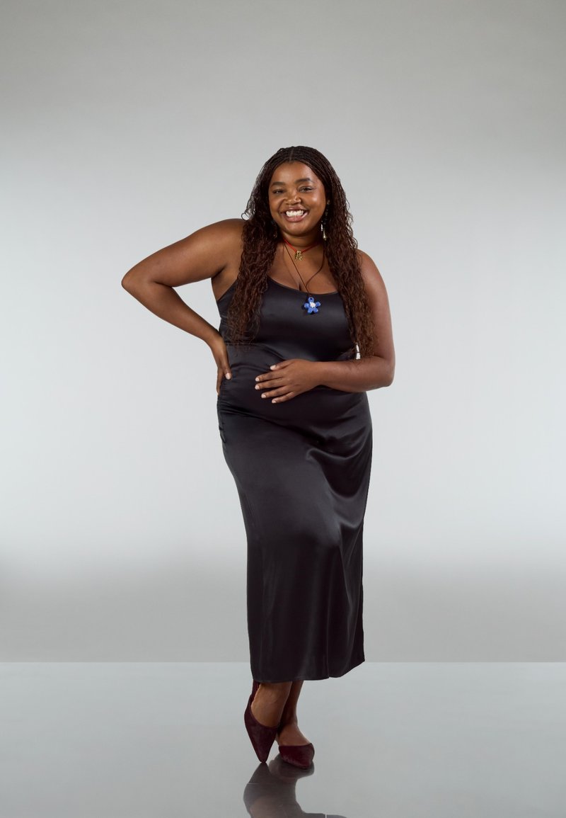 Vestito in raso nero a sottoveste con silhouette aderente. La modella indossa un ciondolo a forma di fiore blu e tacchi color borgogna, in piedi su uno sfondo neutro.