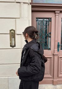 Musta tikattu takki, jossa on huppu, yhdistettynä ruskeaan laukkuun. Kultainen sisäpuhelin vaalealla seinällä, pastellin vaaleanpunainen ovi, jossa koristeellista rautatyötä.