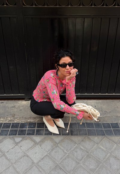 Mujer con cabello rizado oscuro, gafas de sol negras, top rosa con estampado, pantalones negros, zapatos blancos puntiagudos, agachada sobre una acera de baldosa sosteniendo una bolsa beige.