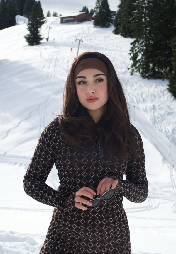 Jeune femme portant un haut à manches longues à motifs et un bandeau, tenant des lunettes de soleil, debout devant une pente enneigée avec des arbres et une cabane en arrière-plan.