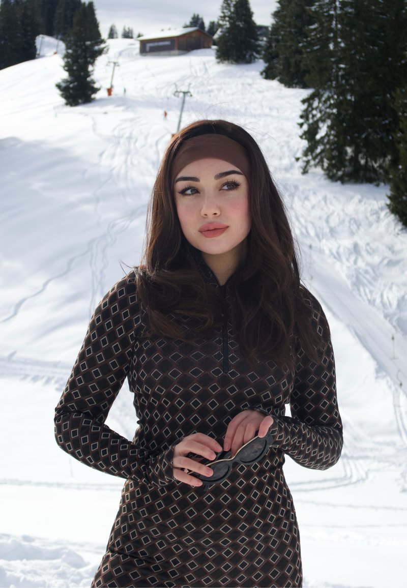 Junge Frau in gemustertem Langarmoberteil und Stirnband hält eine Sonnenbrille, steht vor einem verschneiten Hang mit Bäumen und einer Hütte im Hintergrund.