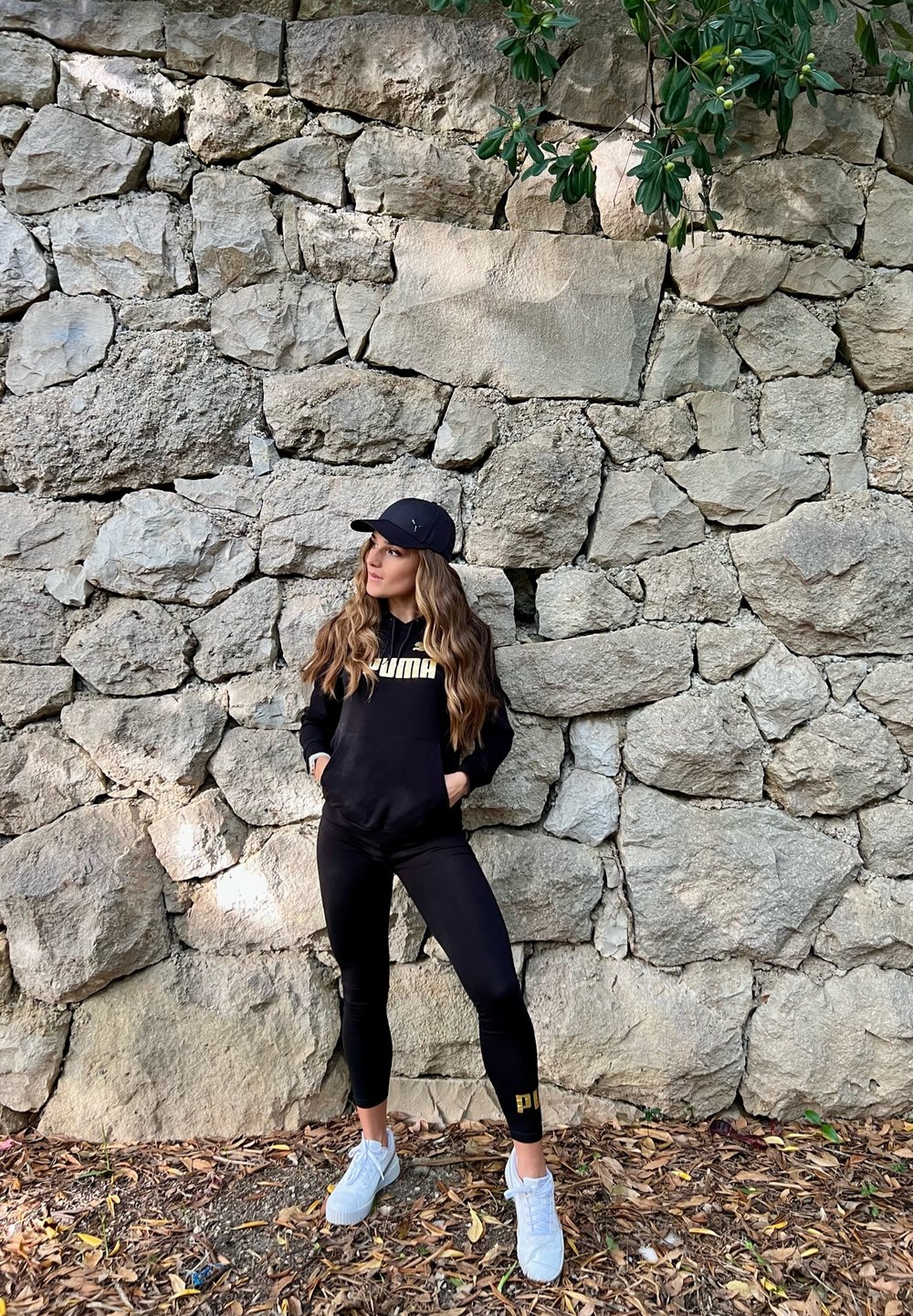 Sudadera negra con el logo "PUMA", leggins negros y zapatillas blancas. De pie contra una pared de piedra texturizada con hojas al fondo.