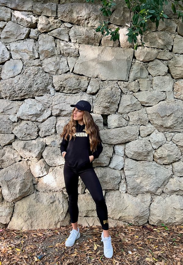 Sudadera negra con el logo "PUMA", leggins negros y zapatillas blancas. De pie contra una pared de piedra texturizada con hojas al fondo.