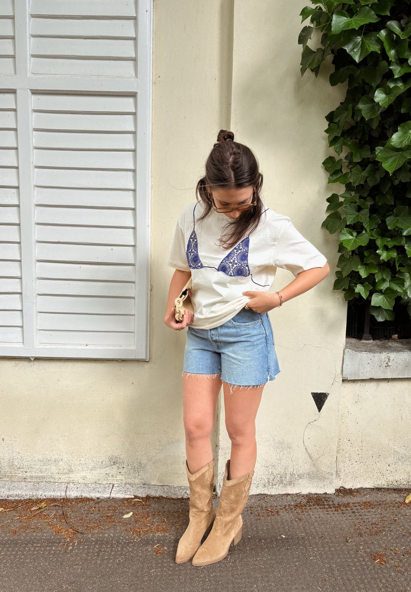 Frau trägt ein weißes T-Shirt mit blauem BH-Aufdruck, Jeans-Shorts, hellbraune Stiefel, hält eine hellfarbige Clutch und steht nahe einer beigen Wand und grünen Blättern.