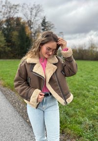 Bruine jas met schapenvachtvoering en een cropped ontwerp, met een grote kraag, met rits afgesloten zakken en een contrasterende beige rand. Lichtblauwe spijkerbroek.