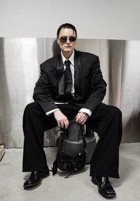 Costume noir avec pantalon large et revers en satin, associé à une chemise blanche et une cravate noire. Des lunettes de soleil noires et un petit sac à main noir complètent le look.