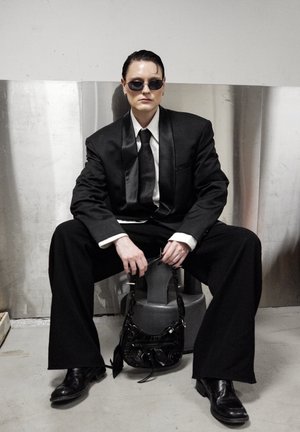 Traje negro con pantalones anchos y solapa de satén, combinado con una camisa blanca y corbata negra. Gafas de sol negras y un pequeño bolso negro completan el conjunto.