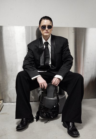 Traje negro con pantalones anchos y solapa de satén, combinado con una camisa blanca y corbata negra. Gafas de sol negras y un pequeño bolso negro completan el conjunto.