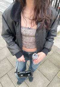 Μαύρο bomber μπουφάν με αφράτο γιακά, λεοπάρ crop top, ψηλόμεσα τζιν, κρατώντας ένα κουτάκι και μια μικρή μαύρη τσάντα.