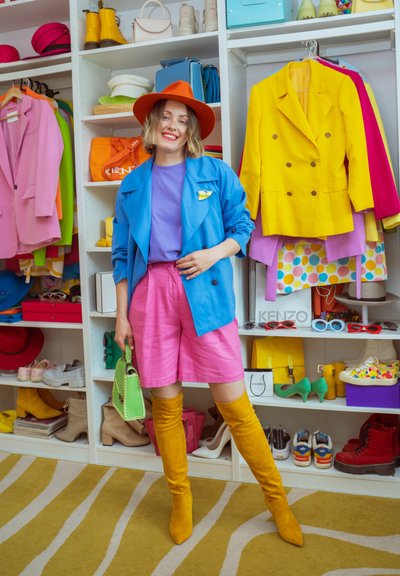 Mujer con chaqueta azul brillante, pantalones cortos rosas, sombrero naranja y botas amarillas hasta la rodilla posa en un armario colorido con zapatos y chaquetas.