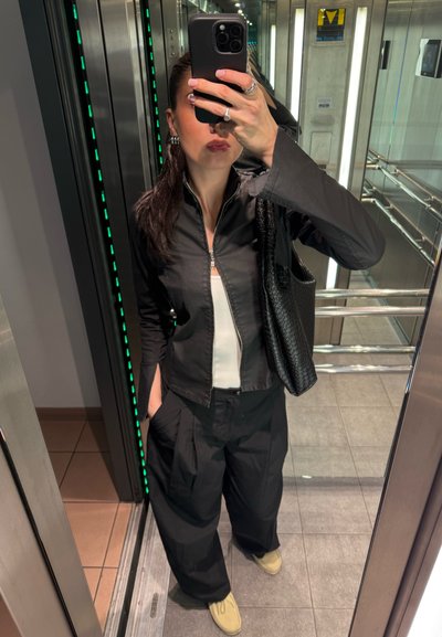 Chaqueta negra con cremallera sobre una blusa blanca, combinada con pantalones holgados negros y zapatos beige. Incluye un bolso negro texturizado. Reflejos en el espejo visibles.