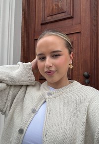Sweter w kolorze białym, wykonany z dzianiny, z okrągłym dekoltem, prążkowanymi mankietami i zapięciem na guziki. Nosi się go na białej bluzce, w połączeniu ze złotymi kolczykami w kształcie serca.