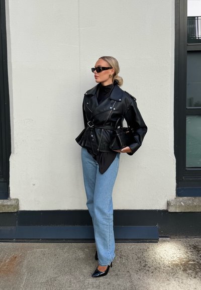 Mujer vestida con chaqueta de cuero negra con cinturón, camiseta de cuello alto negra, jeans azules, tacones negros, gafas de sol y llevando bolso negro, de pie junto a una pared blanca.
