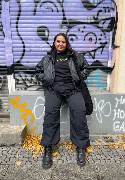 Chaqueta bomber de cuero negro sobre una camiseta negra de marca, combinada con pantalones sueltos negros y botas de cordones negras, con un fondo de graffiti.
