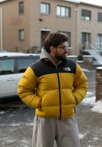 Doudoune jaune avec épaules noires, dotée d'une fermeture éclair à l'avant et d'une capuche. Portée avec un pantalon de survêtement gris, des lunettes visibles. Sol enneigé.