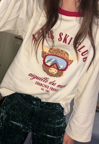 Personne portant une chemise blanche à manches longues avec un buffle en lunettes de ski et le texte rouge « Aries Ski Club, Chamonix France » ainsi qu'un pantalon en velours vert.