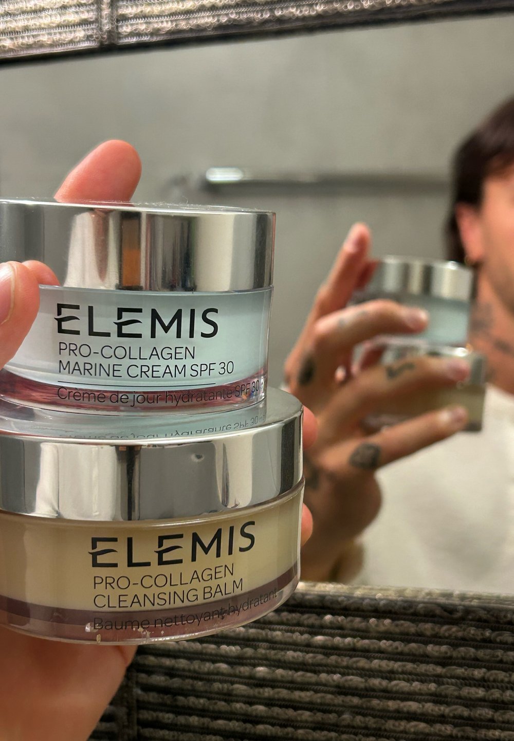 Ruka drži dva ELEMIS jela za njegu kože—Pro-Collagen Marine Cream SPF 30 i Pro-Collagen Cleansing Balm—odbijena u ogledalu kupaonice.