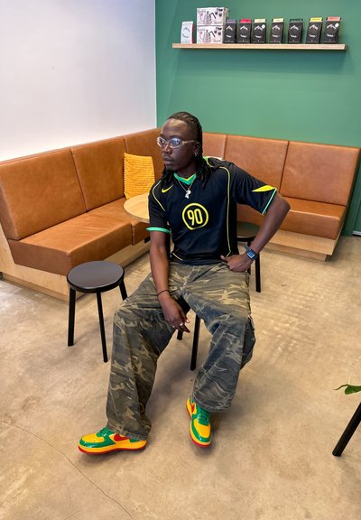 Camiseta de fútbol negra con acentos amarillos, pantalones de camuflaje y zapatillas multicolores; sentado en una silla negra en un ambiente de café.