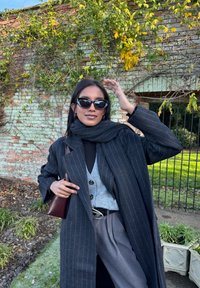 Femme portant un manteau noir à fines rayures, un cardigan gris, un col roulé noir et des lunettes de soleil, posant en extérieur près d'un mur en briques avec des plantes grimpantes.