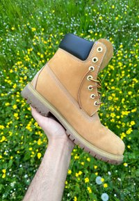 Καφέ μπότες εργασίας με μαύρη κολάρο, χρυσά μάτια και καφέ κορδόνια. Διαθέτουν ανθεκτική σόλα και ραμμένες λεπτομέρειες. Κρατούνται στο χέρι μπροστά από ένα λουλουδάτο φόντο.