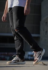 Atletické tenisky v béžové, hnědé a bílé barvě. Mají síťovinovou a semišovou texturu, masivní podešev a výrazné šněrování. Nosené s černými roztrhanými džíny.