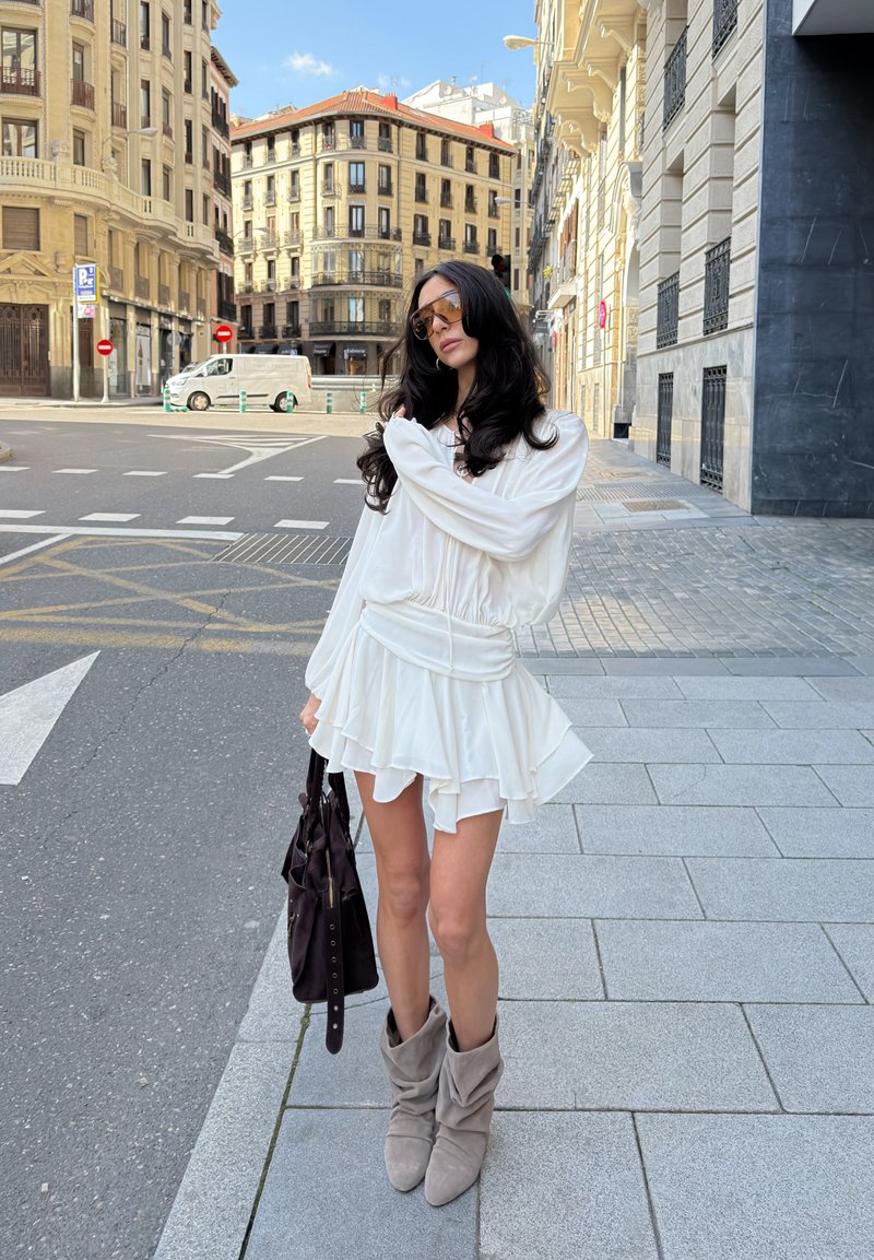 Femme aux longs cheveux foncés portant des lunettes de soleil, une robe blanche, des bottes grises et tenant un sac à main foncé, debout sur un trottoir en ville près d'une intersection.
