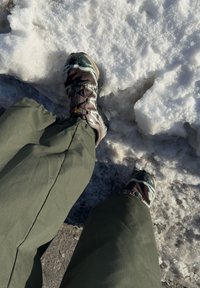 Chaussures brunes et grises avec des semelles texturées, associées à un pantalon vert ample, debout sur de la neige et de la glace.
