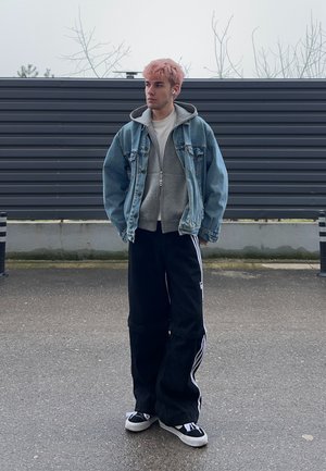 Persona joven con cabello rosa que lleva chaqueta vaquera, sudadera gris con capucha, pantalones anchos negros con rayas blancas y zapatillas negras, de pie al aire libre junto a una cerca.