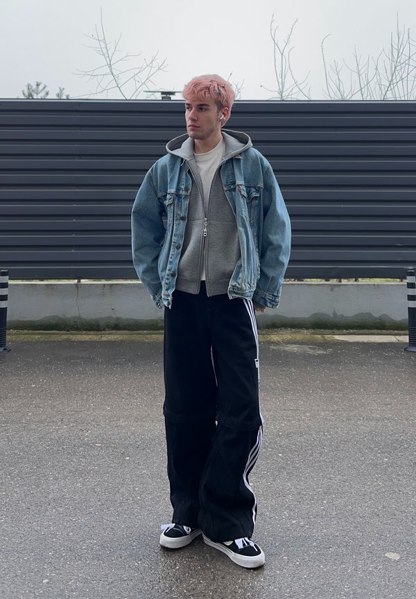 Young person with pink hair wearing a denim jacket, grey hoodie, black wide trousers with white stripes, and black trainers, standing outdoors against a fence.
