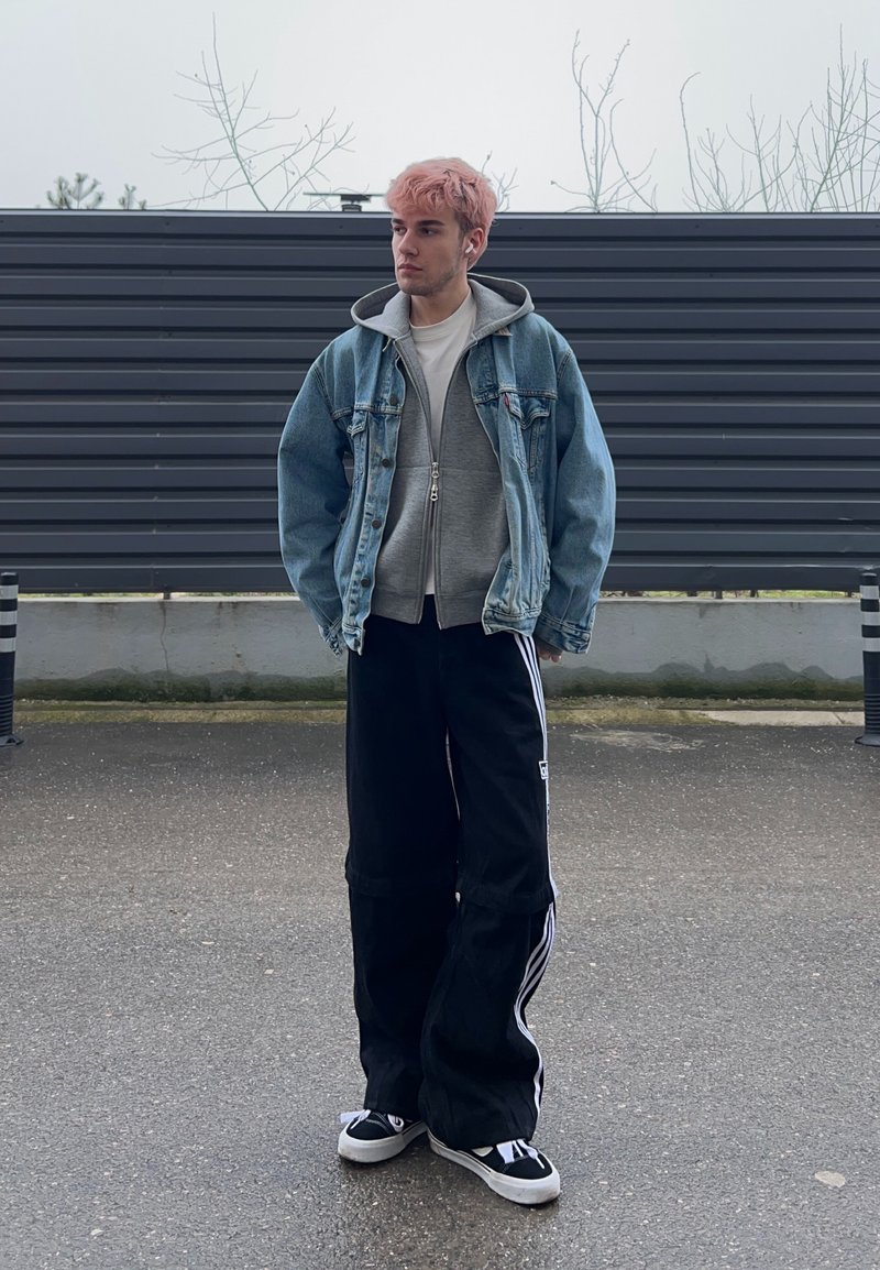 Jeune personne aux cheveux roses portant une veste en jean, un sweat à capuche gris, un large pantalon noir avec des rayures blanches et des baskets noires, debout en plein air contre une clôture.