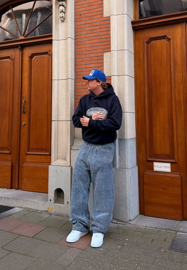 Noor mees sinise nokamütsi, musta kapuutsiga, lahtiste hallide teksade ja valgete tossudega seisab kivi- ja tellismüüri vastu puidust uste vahel.