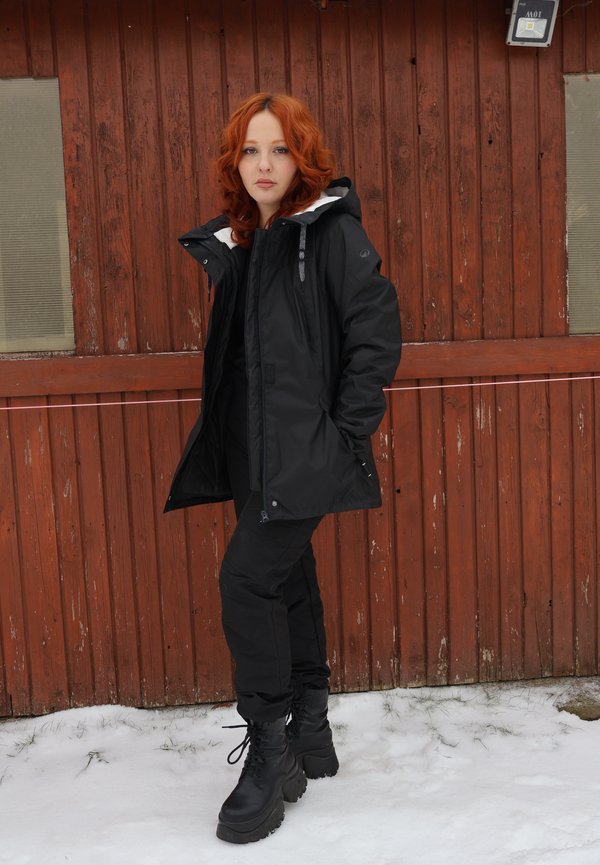 Giovane donna con capelli rossi che indossa una giacca invernale nera, pantaloni e stivali, in piedi sulla neve davanti a un capanno di legno rosso logorato dal tempo.