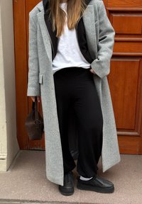 Manteau en laine gris, veste zippée noire, t-shirt blanc, pantalon large noir, chaussures noires sans lacets et petit sac à main marron.