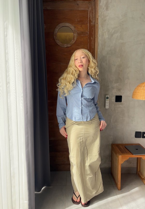 Žena s dlhými blond vlasmi, oblečená v modrej košeli na gombíky a béžovej sukni, stojí pri okne so závesmi v miestnosti s drevenými a betónovými stenami.