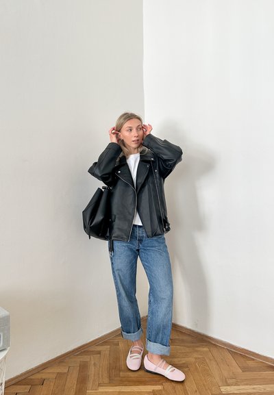 Chaqueta de moto de cuero negro, camiseta blanca, jeans azules remangados, bailarinas rosas y un bolso negro contra una pared clara. Textura de suelo de madera.