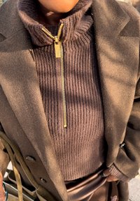 Personne portant un pull marron côtelé avec une fermeture éclair dorée et un manteau marron, portant un sac avec une sangle marron visible.