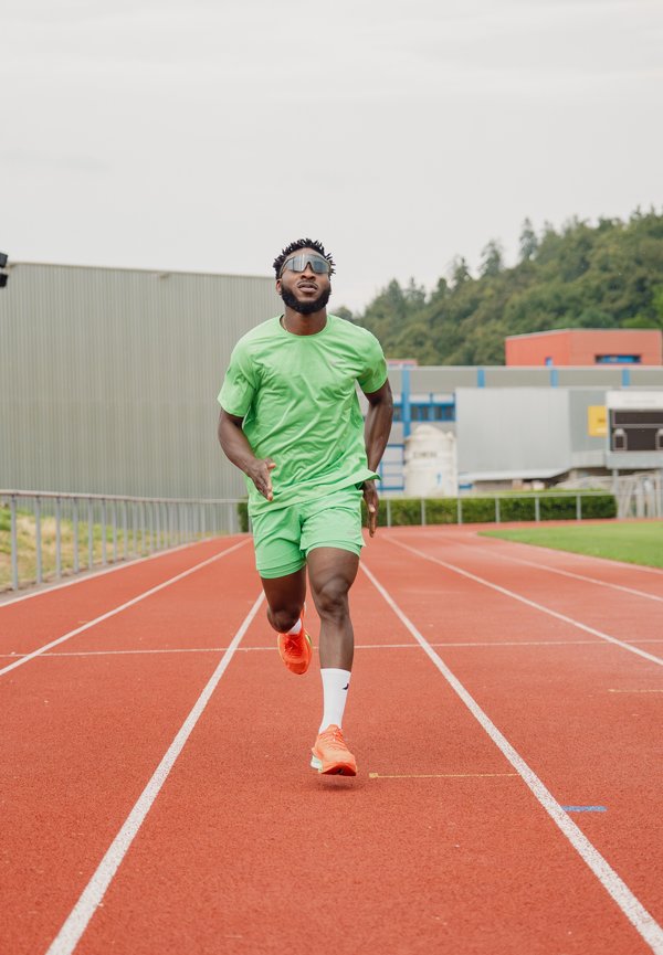 Homme portant des vêtements de sport verts et des chaussures orange courant sur une piste extérieure rouge avec des bâtiments et des arbres en arrière-plan.