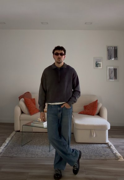 Hombre con gafas de sol, suéter oscuro y vaqueros, de pie con una mano en el bolsillo en una sala de estar moderna con sofá beige y cojines rojos.