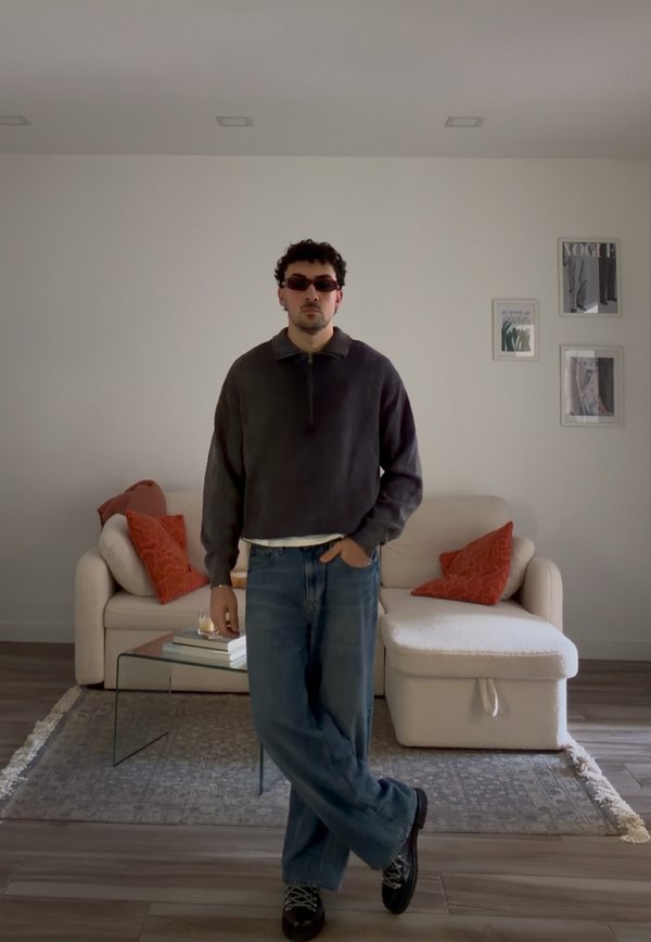 Man met zonnebril, donker trui en spijkerbroek staat met één hand in zak in een moderne woonkamer met beige bank en rode kussens.