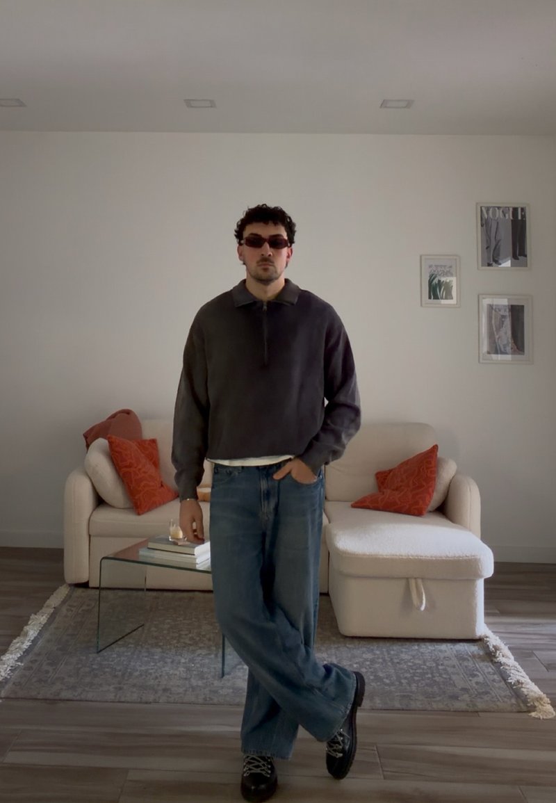 Mand iført solbriller, mørk sweater og jeans står med den ene hånd i lommen i en moderne stue med beige sofa og røde puder.