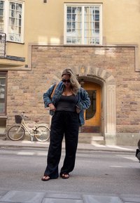 Τζάκετ τζιν, μαύρο καλσόν, και παντελόνι φαρδύ. Σανδάλια, κυματιστά ξανθά μαλλιά, στέκεται μπροστά από ένα πέτρινο κτίριο με ξύλινη πόρτα, ποδήλατο κοντά.