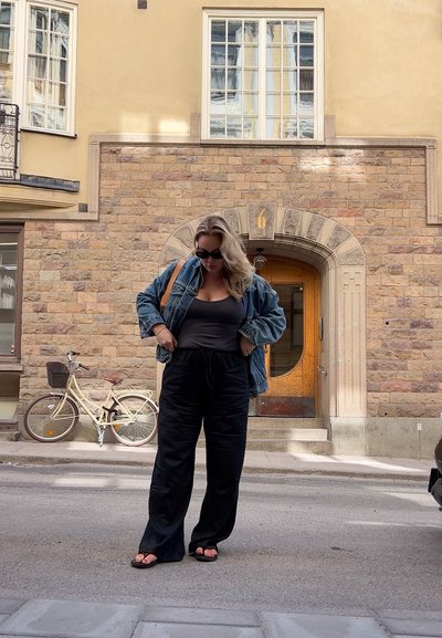 Chaqueta de mezclilla, top negro sin mangas y pantalones de pierna ancha. Sandalias, cabello rubio ondulado, de pie frente a un edificio de piedra con puerta de madera, bicicleta cerca.