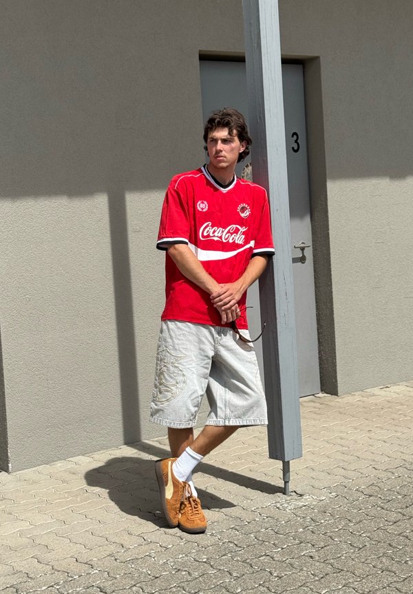 Giovane uomo con maglietta rossa Coca-Cola e pantaloncini chiari si appoggia a un palo grigio vicino a una porta grigia con il numero tre.