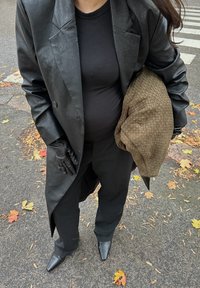 Casaco longo de couro preto sobre uma blusa preta ajustada, combinado com calças escuras e botins pretos de bico fino. Bolsa texturizada castanha debaixo do braço.
