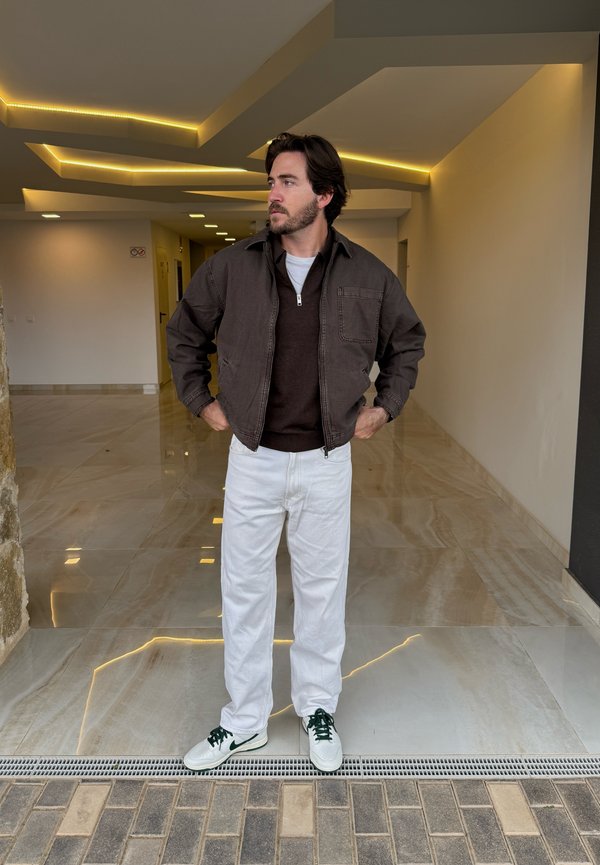 Veste marron par-dessus un pull marron foncé, pantalons blancs et baskets blanches avec des accents verts. Cadre intérieur avec des sols en marbre et un éclairage indirect.