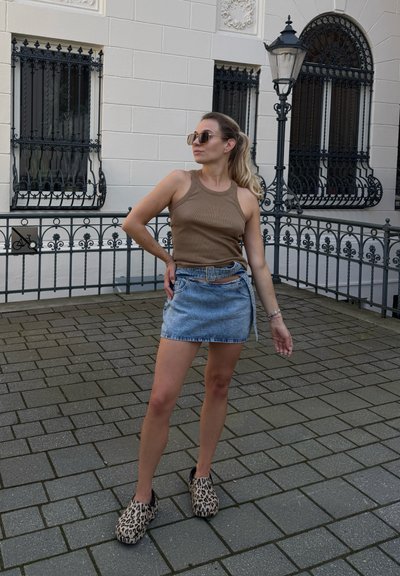Camiseta sin mangas marrón de canalé, falda de mezclilla azul claro con cintura con cinturón y zuecos de estampado de leopardo, posando sobre una superficie pavimentada cerca de ventanas decorativas.