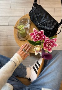 Persoon in jeans en witte trui die uitstrekt naar een houten tafel met een ijskoud groen drankje, een glas water, roze bloemen en een zwarte tas.