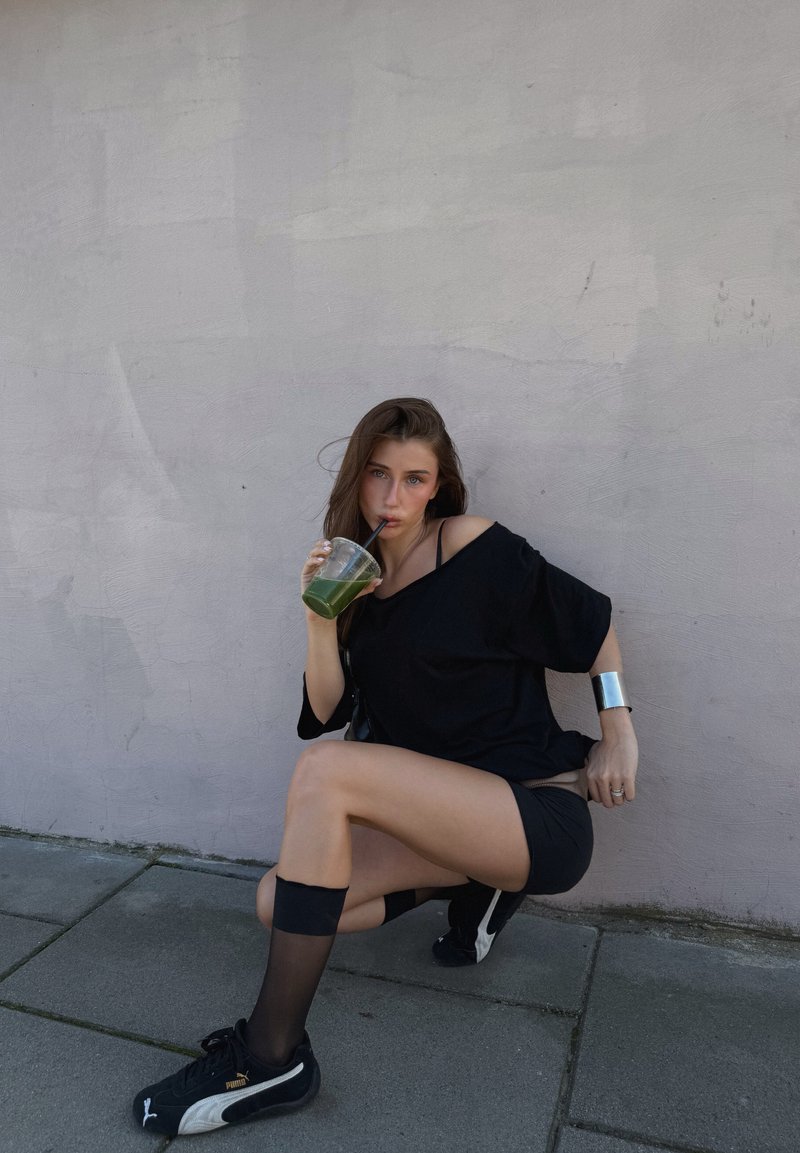 Modello in una maglietta nera oversize e pantaloni corti, che tiene in mano un bicchiere trasparente con una bevanda verde, indossando scarpe da ginnastica nere e un bracciale in argento.